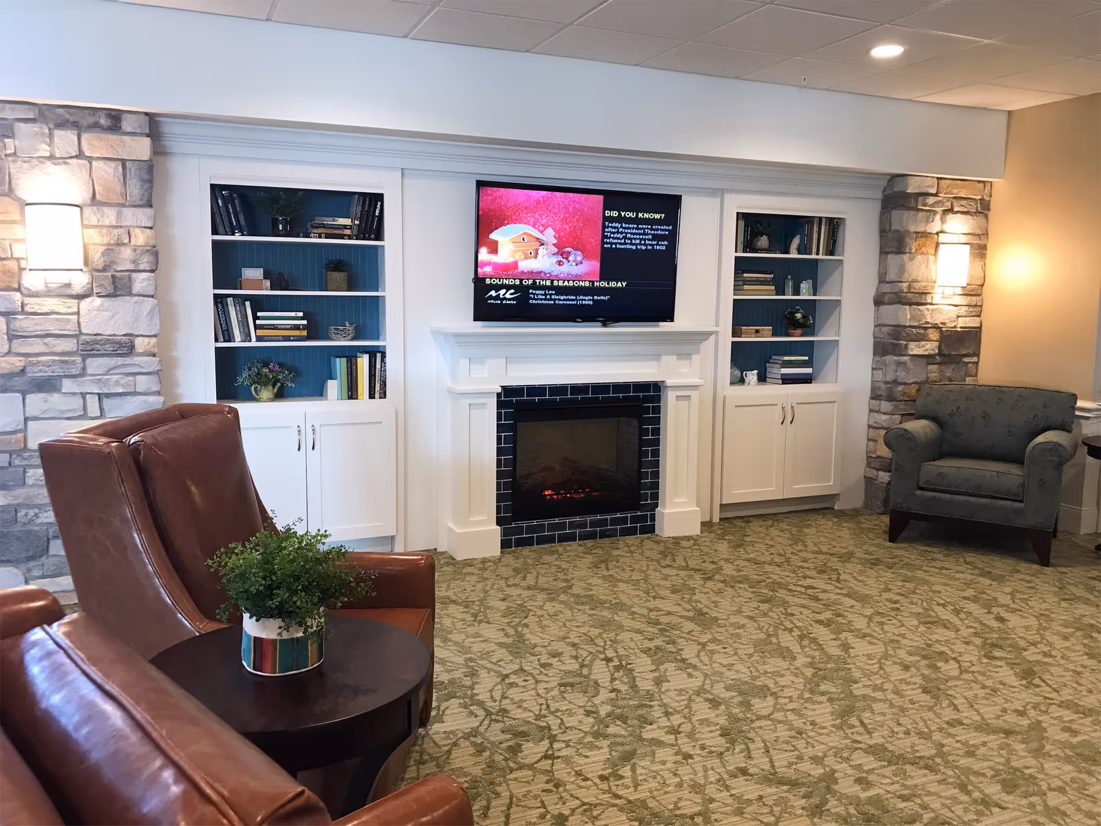 Cozy common area with a fireplace and TV flanked by built-in bookshelves, leather and upholstered armchairs, and a side table with a plant.