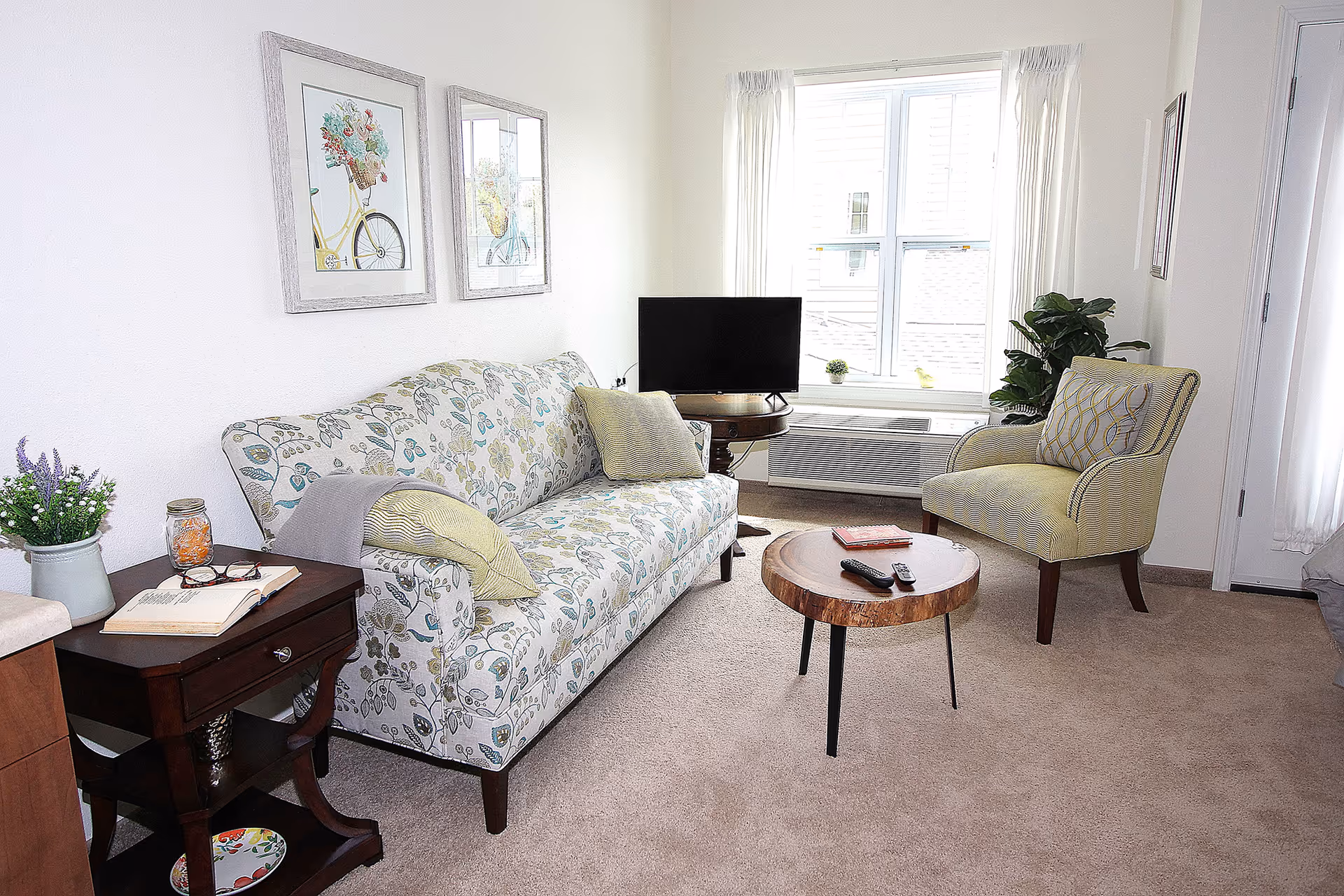 Bright living room with a patterned sofa, armchair, round wooden coffee table, TV on a stand, and a window letting in natural light.