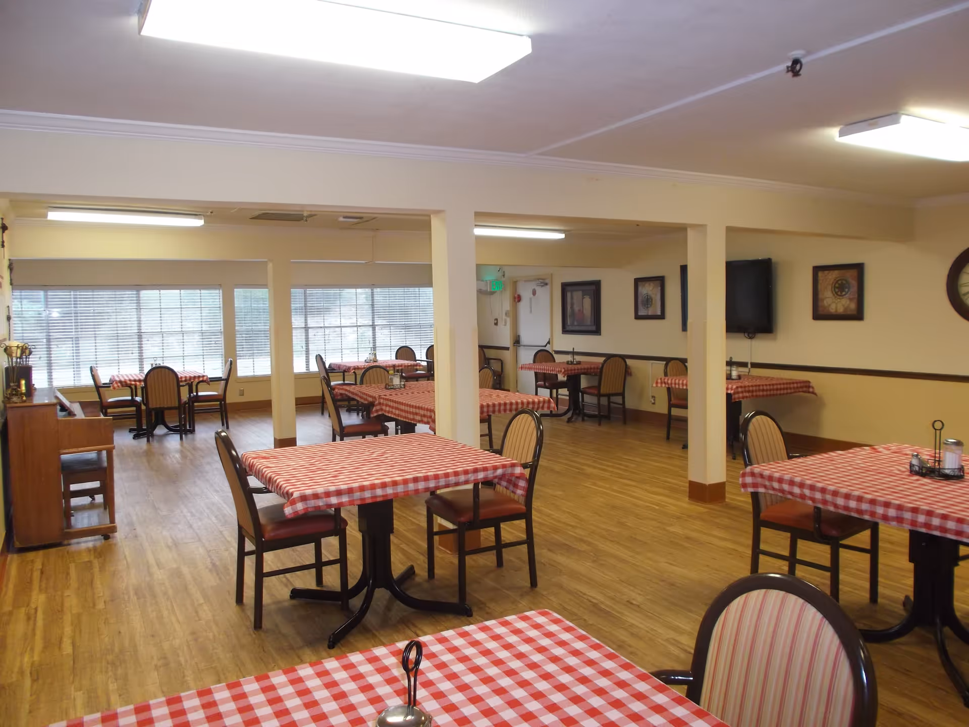 Interior view of a dining room in a senior living facility with multiple tables covered in red and white checkered tablecloths, surrounded by chairs. The room has wooden flooring, large windows with blinds, framed artwork on the walls, and ceiling lights.