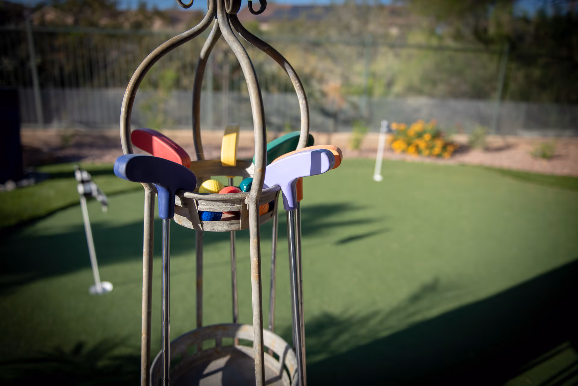Close-up view of a metal golf club holder containing colorful mini golf putters and balls on a putting green with a flagstick and hole in the background, surrounded by a garden and fencing.