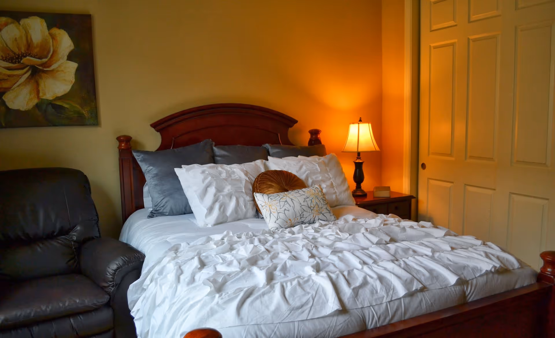 A cozy bedroom with a wooden bed frame, white ruffled bedding, multiple pillows including decorative ones, a bedside table with a lit lamp, a dark leather armchair, a floral painting on the wall, and a closed closet with sliding doors.