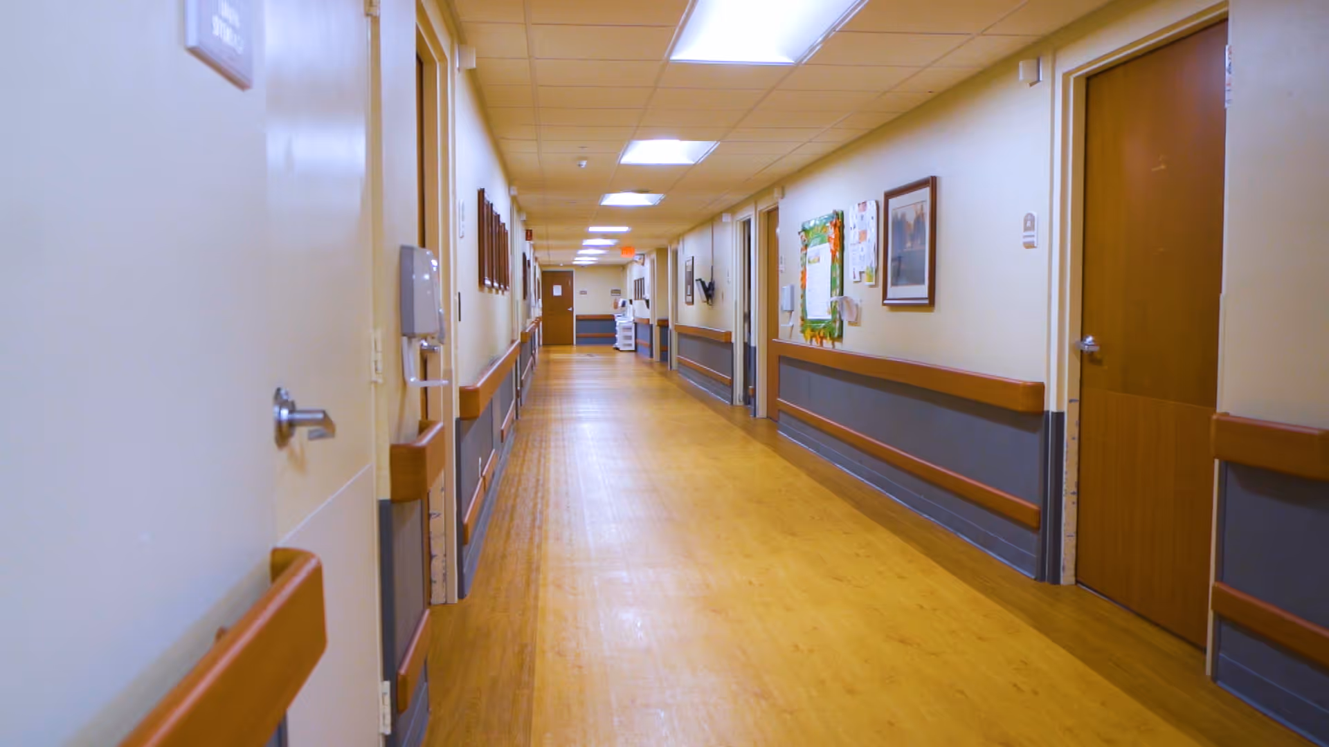 A long, well-lit hallway in a senior living facility with wooden handrails on both sides, light-colored walls, several closed doors, framed pictures, and a bulletin board on the right wall.