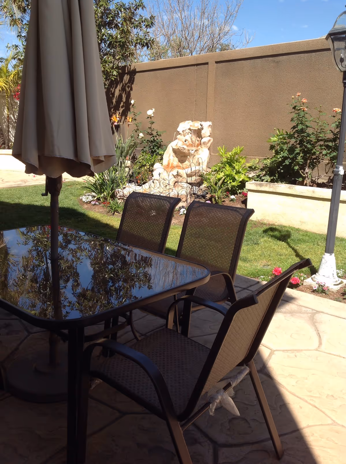 Outdoor patio area with a glass-top table and four brown mesh chairs. A closed beige umbrella is positioned in the center of the table. In the background, there is a garden with various plants, flowers, a stone water fountain, and a tall brown wall. A lamp post is visible on the right side.