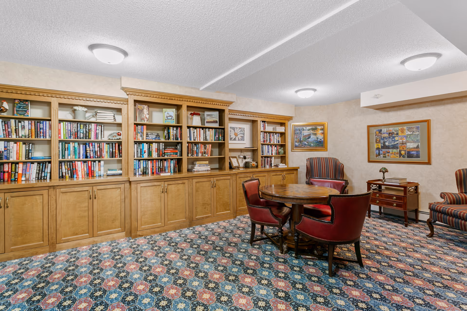 A cozy library or reading room with wooden bookshelves filled with books along the back wall. In front of the shelves is a round wooden table surrounded by four red leather chairs. The room has patterned carpet flooring, beige walls with framed artwork, and two striped armchairs near a small wooden side table with a lamp.