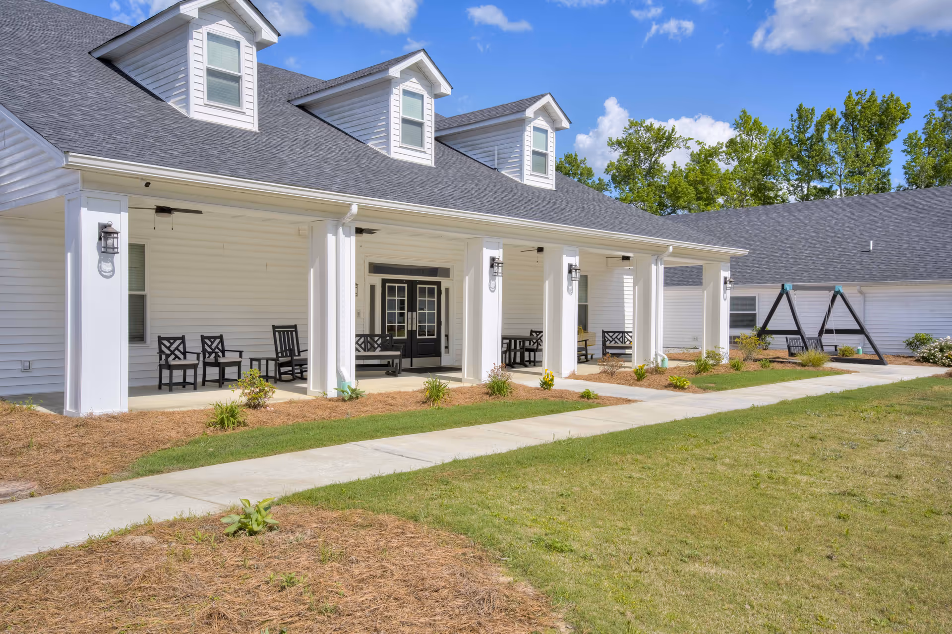 Exterior view of a white senior living facility building with a covered porch featuring several black chairs and benches. The building has a gray shingled roof with dormer windows, and there is a paved walkway leading up to the porch. The surrounding area has grass, small plants, and trees in the background under a blue sky with some clouds.
