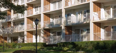 Exterior view of a multi-story residential building with balconies, brick facade, white railings, lamp posts, and landscaped grounds.
