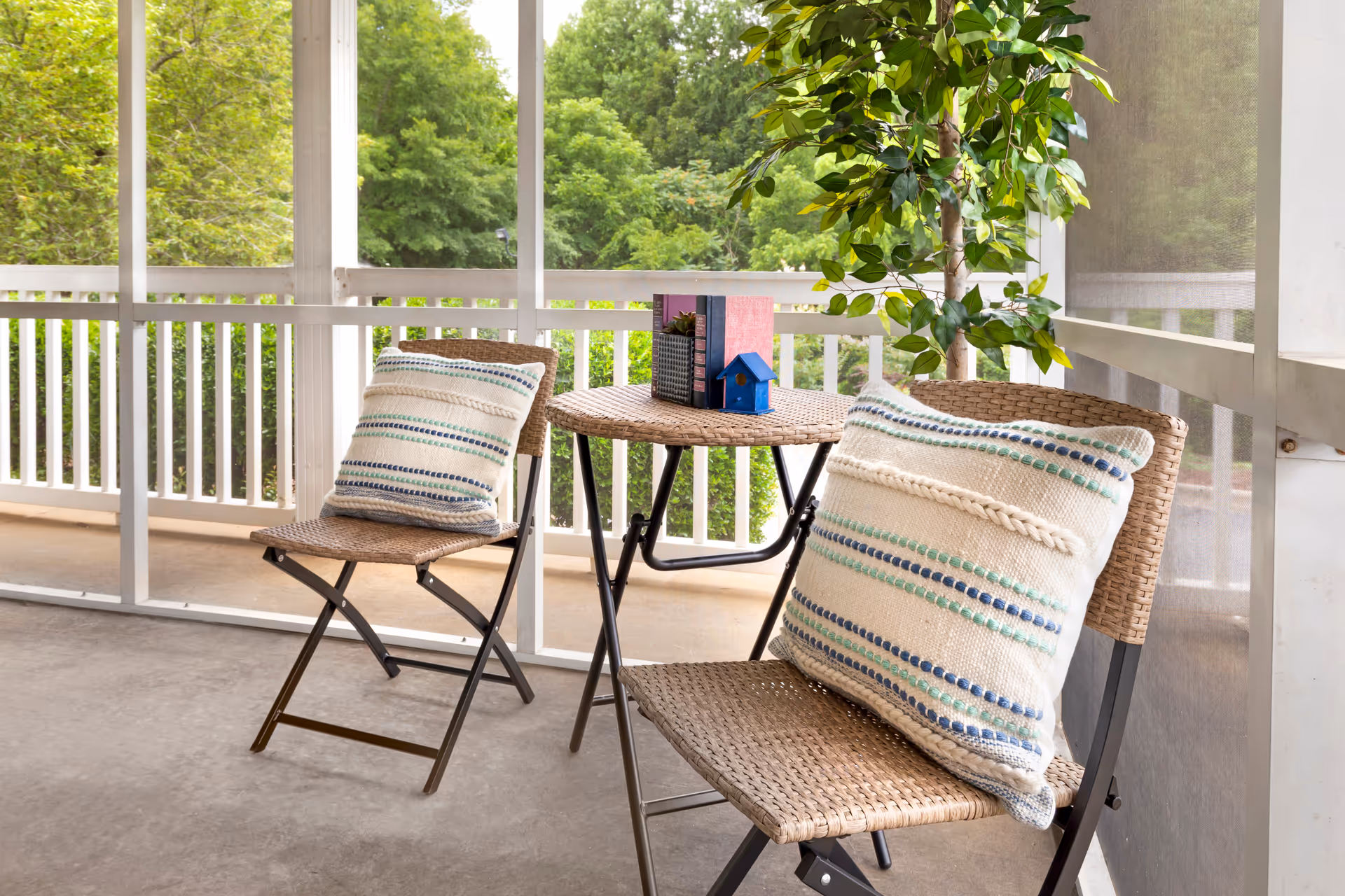 A cozy screened-in porch with two wicker chairs, each with a striped cushion, and a small round wicker table between them. On the table are two books, a small potted plant, and a miniature blue birdhouse. Lush green trees are visible outside the porch.