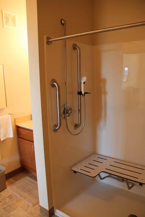Accessible bathroom shower with grab bars, a handheld shower head and a fold-down bench next to a vanity.