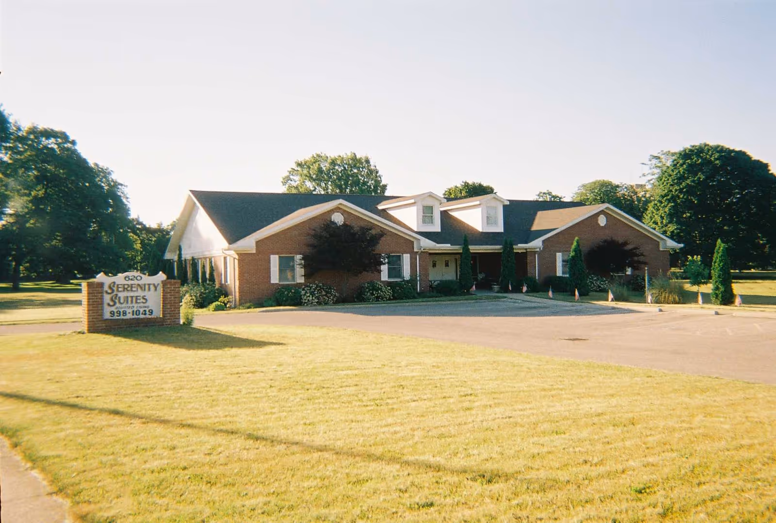 Exterior view of a single-story brick building with a dark roof, surrounded by a well-maintained lawn and trees. A sign in front reads '620 Serenity Suites 998-1049'. The sky is clear and the area appears calm and sunny.