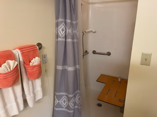 Bathroom with a shower area featuring a gray patterned shower curtain, a wooden fold-down shower seat, and grab bars. On the left wall, there are two white towels with red towels folded and placed on top, hanging on a towel rack. A light switch is visible on the right wall.