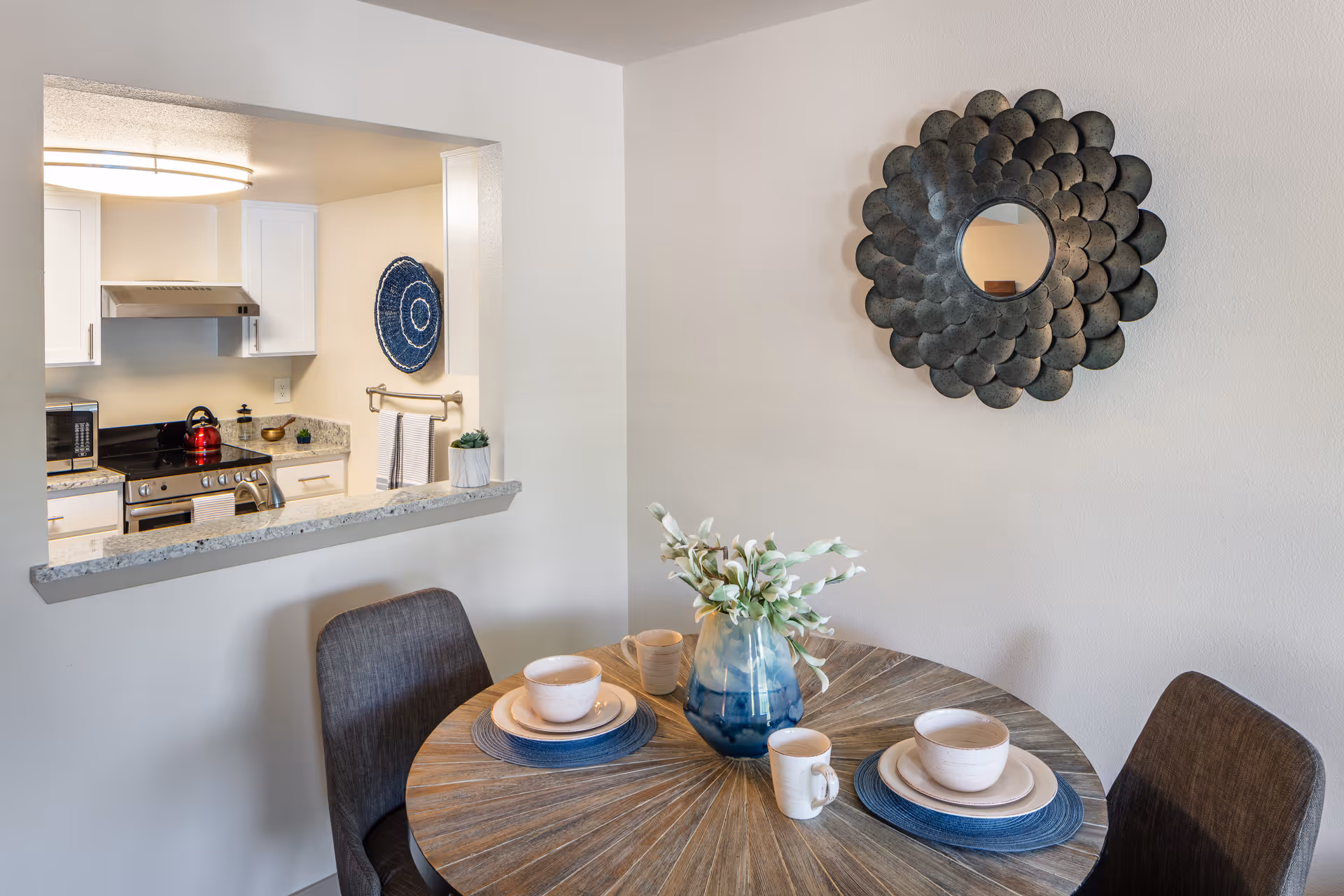 Small dining area with a round wooden table set for two, a blue vase centerpiece and decorative wall mirror, seen beside a pass-through window into a white kitchen.