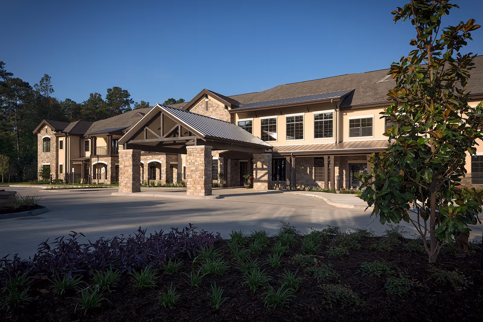 Exterior view of a two-story senior living facility building with a covered entrance supported by stone pillars, surrounded by landscaped greenery and trees under a clear blue sky.