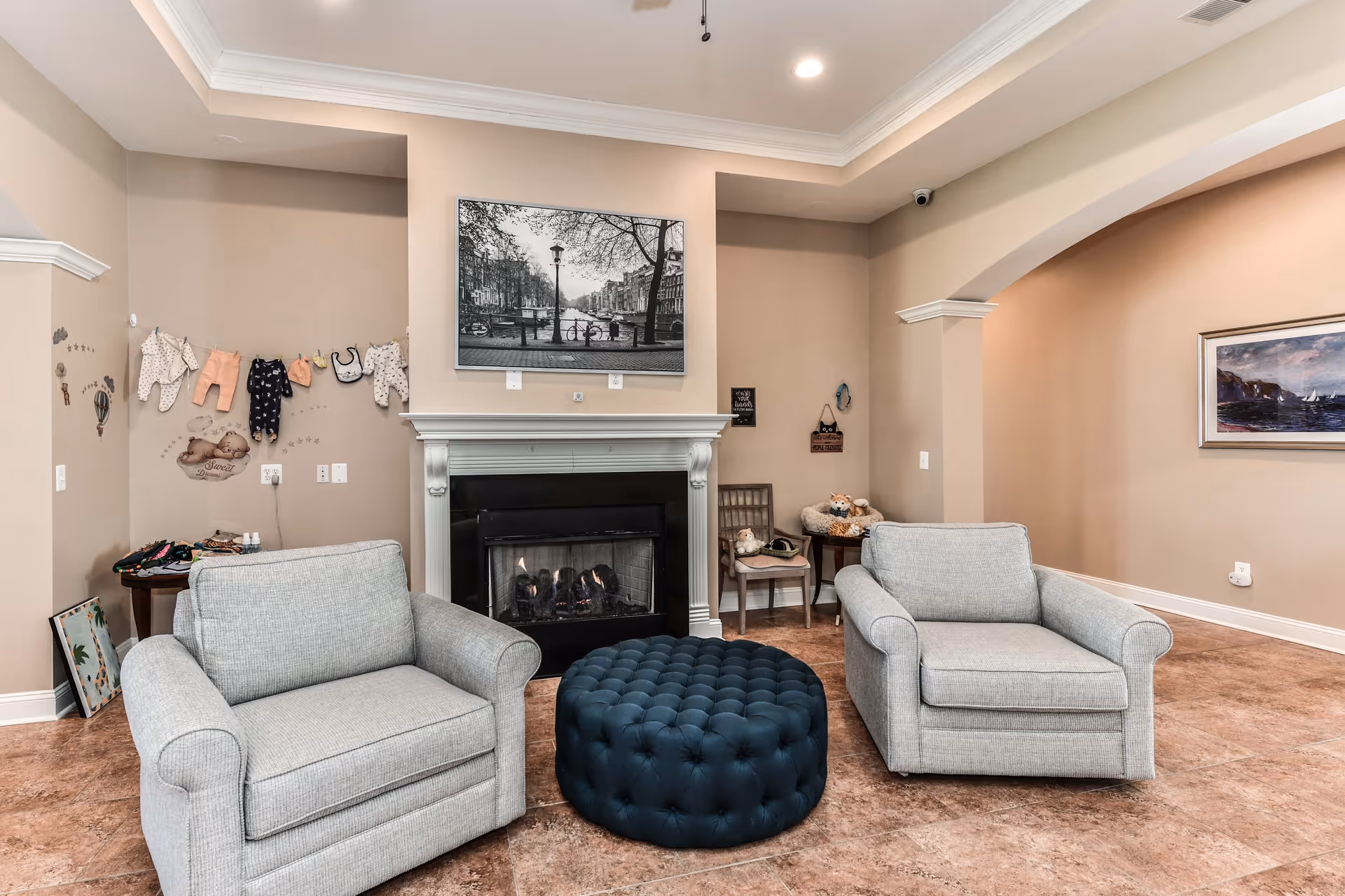 A cozy living room area with two light gray armchairs facing a black fireplace with a white mantel. Above the fireplace is a black and white framed photograph of a street scene. Between the chairs is a round, tufted dark blue ottoman. The walls are painted beige, and there are decorative items including baby clothes hanging on the left wall and a chair with stuffed animals on the right side. The floor is tiled in a warm brown tone.