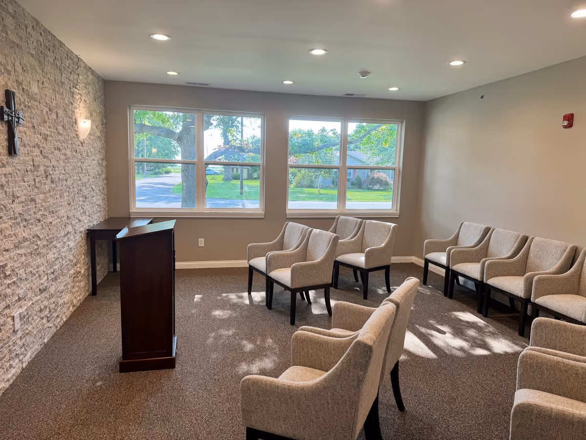 A small meeting or chapel room with beige upholstered chairs arranged in rows facing a wooden podium. The room has a stone accent wall with a cross mounted on it, beige walls, and large windows letting in natural light with a view of green trees and a lawn outside.