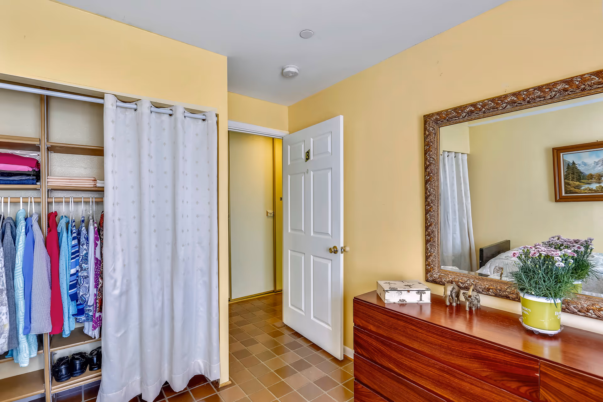 Interior view of a bedroom with a wooden dresser topped with a potted plant, decorative box, and small elephant figurines. A large ornate mirror hangs above the dresser reflecting part of the bed and a framed landscape painting. To the left, there is an open closet with clothes hanging and folded on shelves, partially covered by a light-colored curtain. The room has yellow walls and a tiled floor, with a white door slightly open showing a hallway.