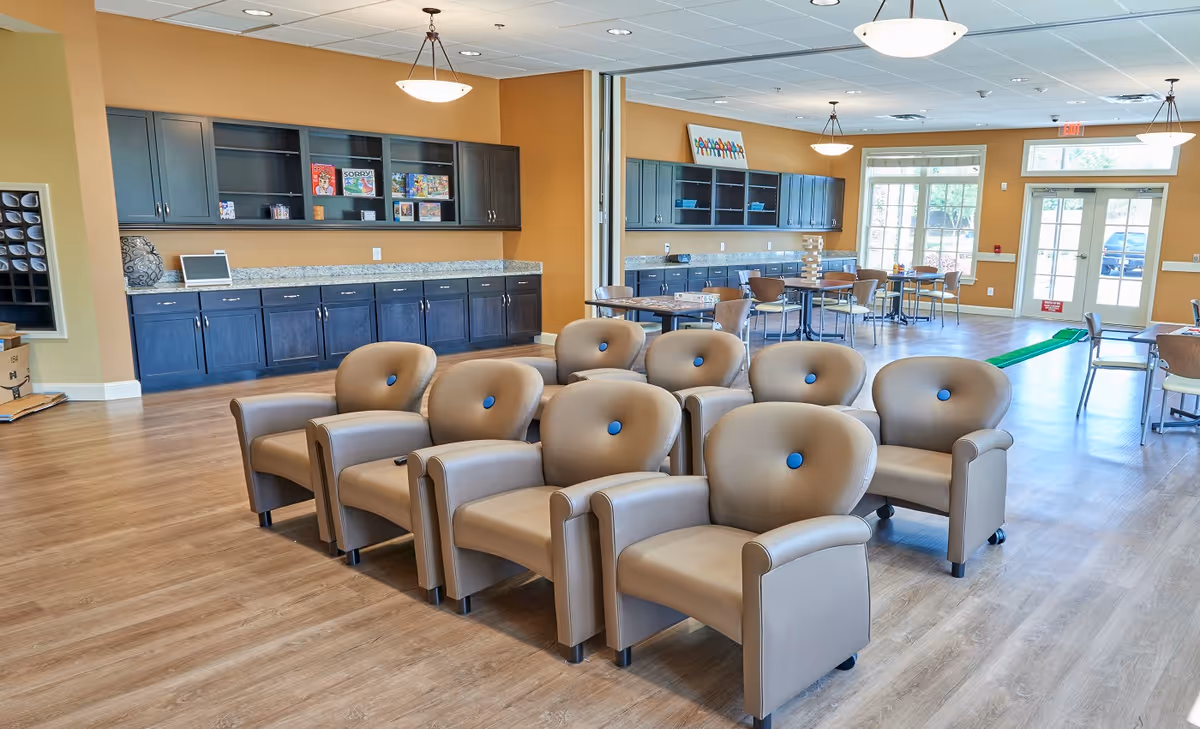 A spacious common area with tan leather armchairs arranged in rows facing forward. Behind the chairs, there are tables and chairs for group activities or dining. The walls are painted a warm beige color, and there are dark wood cabinets with shelves holding board games and other items. Large windows and glass doors allow natural light to fill the room.