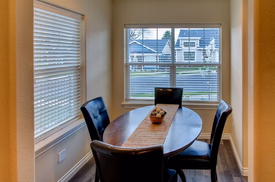 A small dining area with an oval wooden table and four black leather chairs. The table has a woven runner and a decorative bowl with natural elements. Two windows with white blinds allow natural light to enter, showing a view of houses and greenery outside.