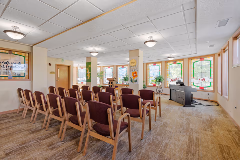 A bright room with rows of wooden chairs with maroon cushions arranged facing a podium and an electronic keyboard. The room has large stained glass windows with words like 'Peace', 'Hope', and 'Joy' displayed. The walls have inspirational signs and there are plants near the windows.