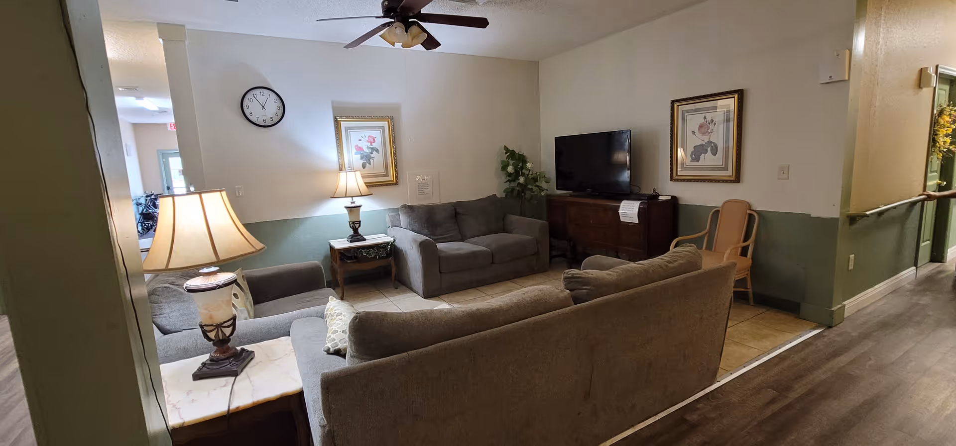 A cozy living room area in a senior living facility with three gray upholstered sofas arranged around a small wooden table with a lamp. A wall clock and framed floral artwork hang on the walls. A flat-screen TV sits on a wooden cabinet, and a wooden chair is placed nearby. The room has tiled flooring and a ceiling fan with lights. The hallway with wood-style flooring is visible to the right.