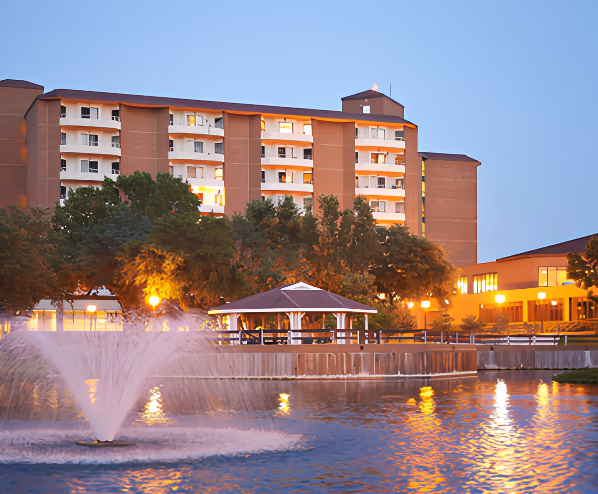 Multi-story senior living building at dusk with illuminated windows, a gazebo, trees, and a fountain in a reflecting pond.