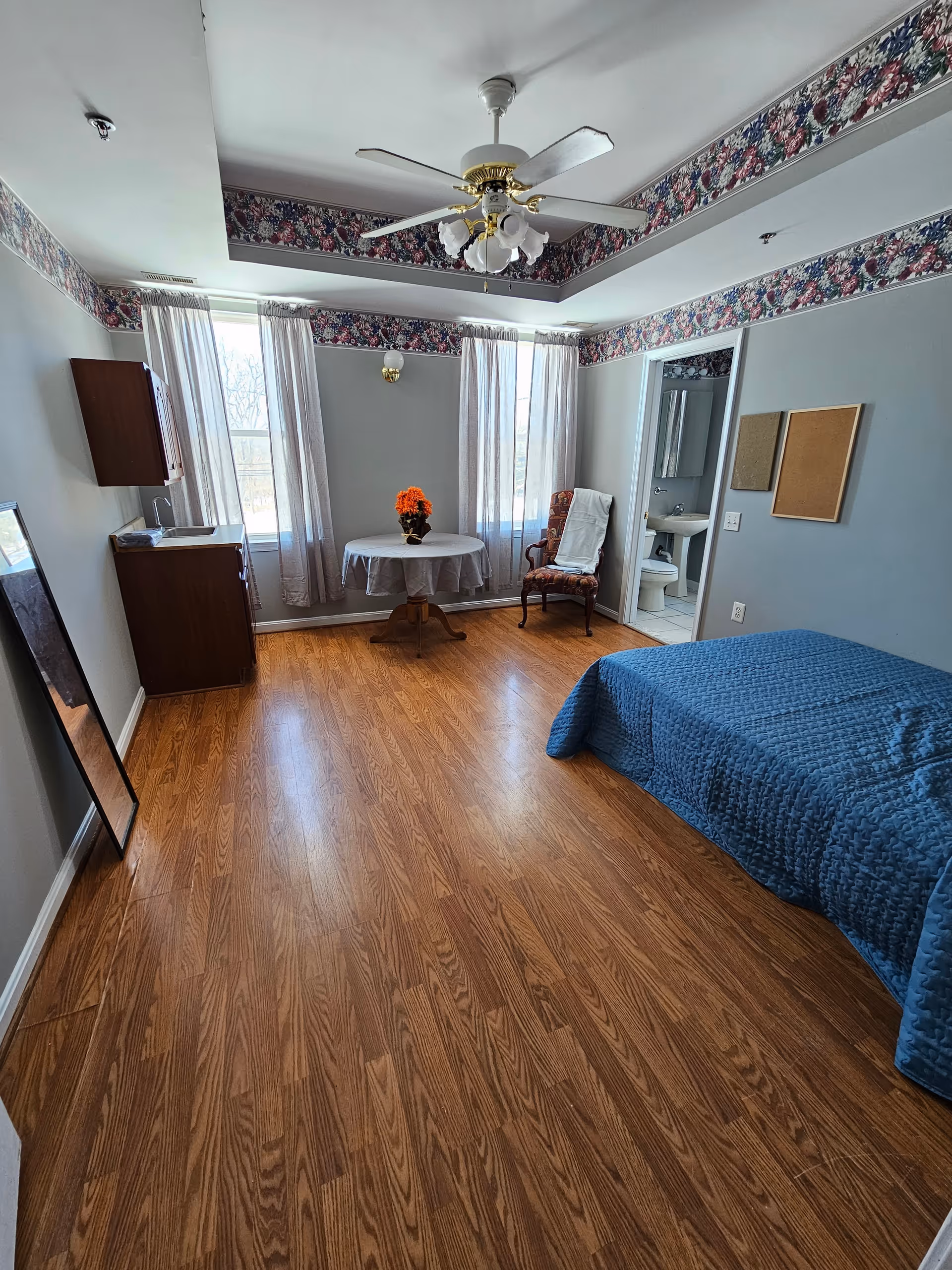 A bright assisted living bedroom with wood flooring, a blue bedspread on a bed, a small round table with a flower vase, an armchair with a white cloth draped over it, two large windows with sheer curtains, a ceiling fan with lights, a wall-mounted cabinet, a full-length mirror leaning against the wall, and an open door leading to a bathroom with a toilet and sink.