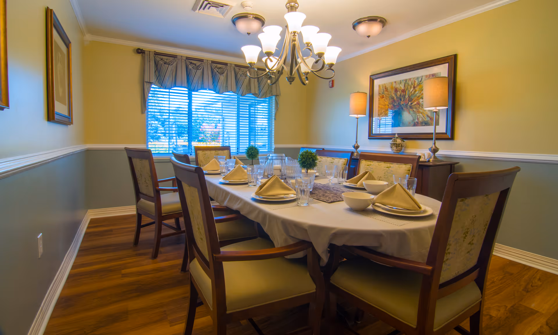 A dining room with a rectangular table set for six people. The table is covered with a white tablecloth and has plates, bowls, glasses, and folded napkins. There are six wooden chairs with cushioned seats around the table. The room has wooden flooring, yellow walls with white trim, a large window with blinds and a valance, two table lamps on a sideboard, and a framed colorful painting on the wall.