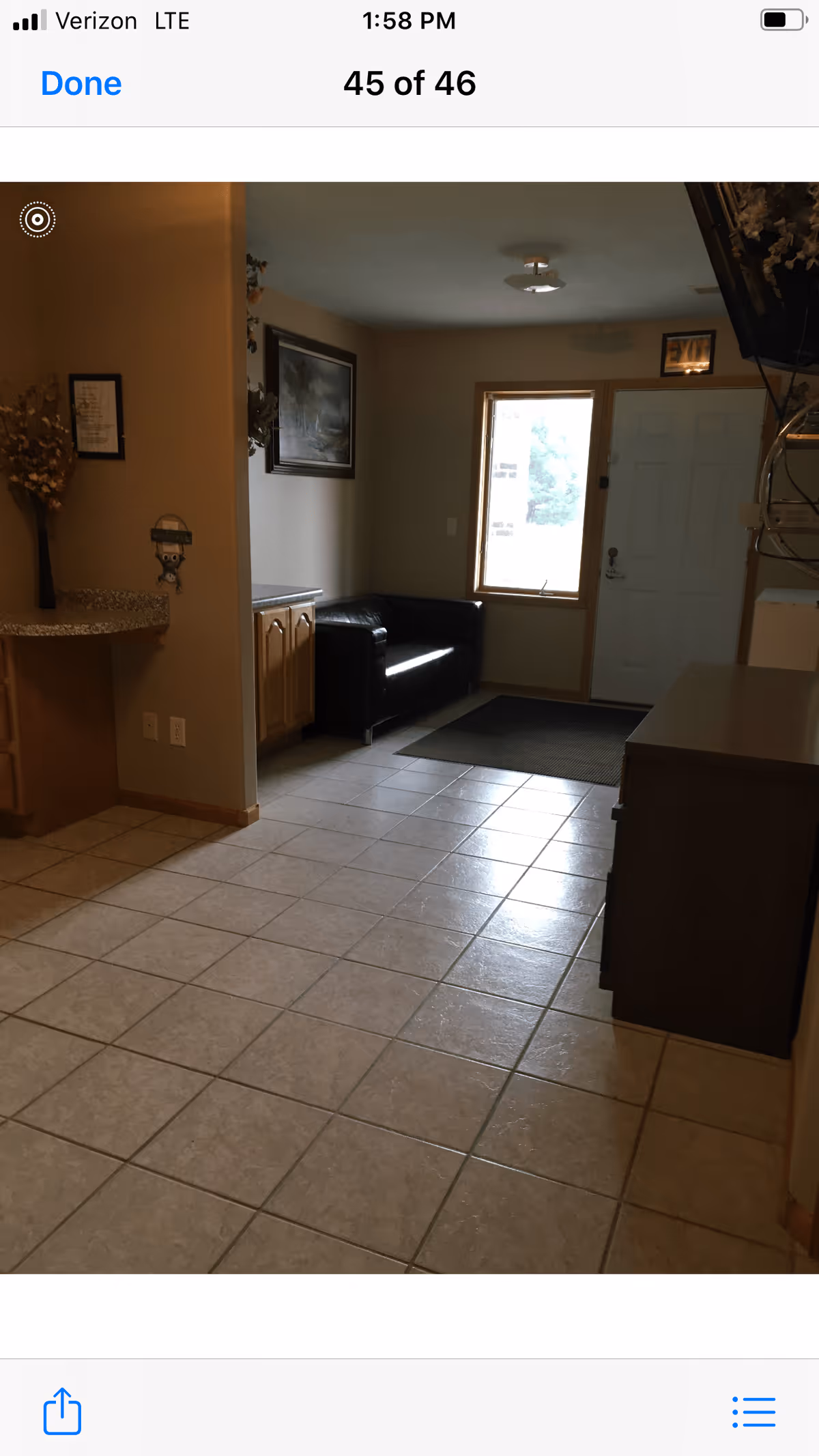 Interior view of a room with tiled floor, a black leather couch near a window and a white door. There is a countertop with a vase of dried flowers on the left side and a cabinet on the right side. The room is softly lit with neutral-colored walls.