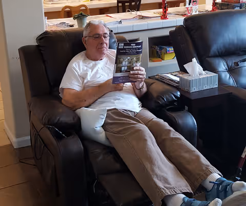 An elderly man wearing glasses, a white t-shirt, and brown pants is sitting comfortably in a dark brown recliner chair, reading a brochure. Next to him is another recliner with a box of tissues on a small table between the chairs. The background shows a kitchen counter with various items on it.