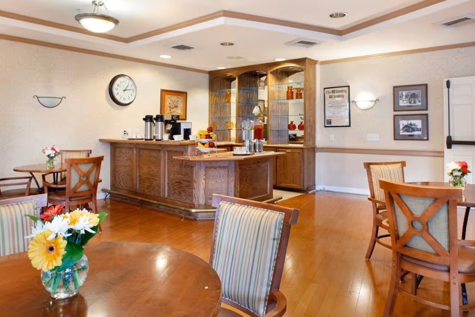 A cozy dining area with wooden tables and chairs, some tables decorated with flower vases. There is a wooden counter with coffee and juice dispensers, a clock on the wall, framed pictures, and soft lighting fixtures on the walls and ceiling.