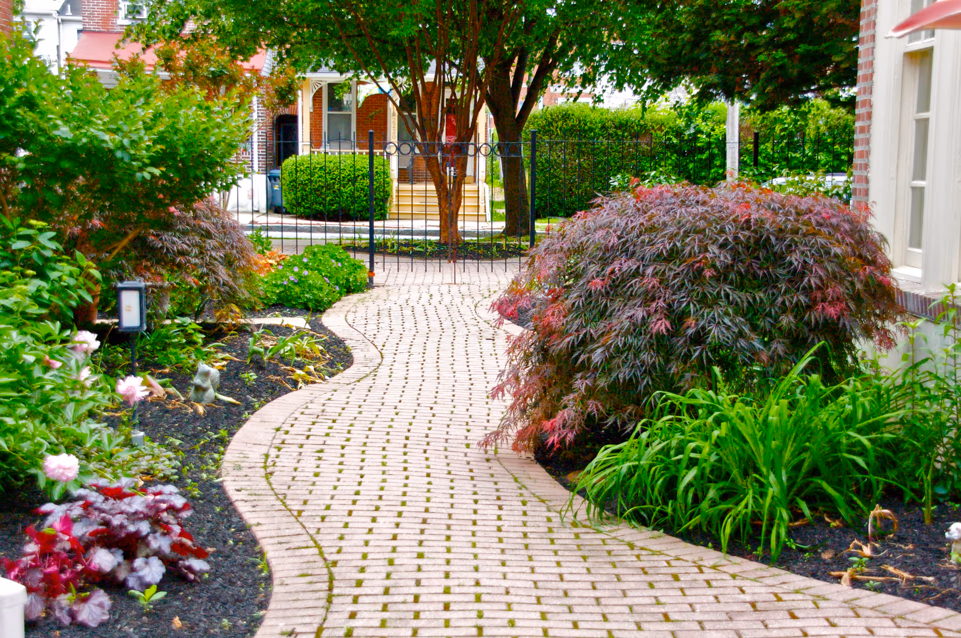 A curved brick pathway lined with various green and red shrubs and plants leading to a black metal gate in front of a brick building with steps and a porch.