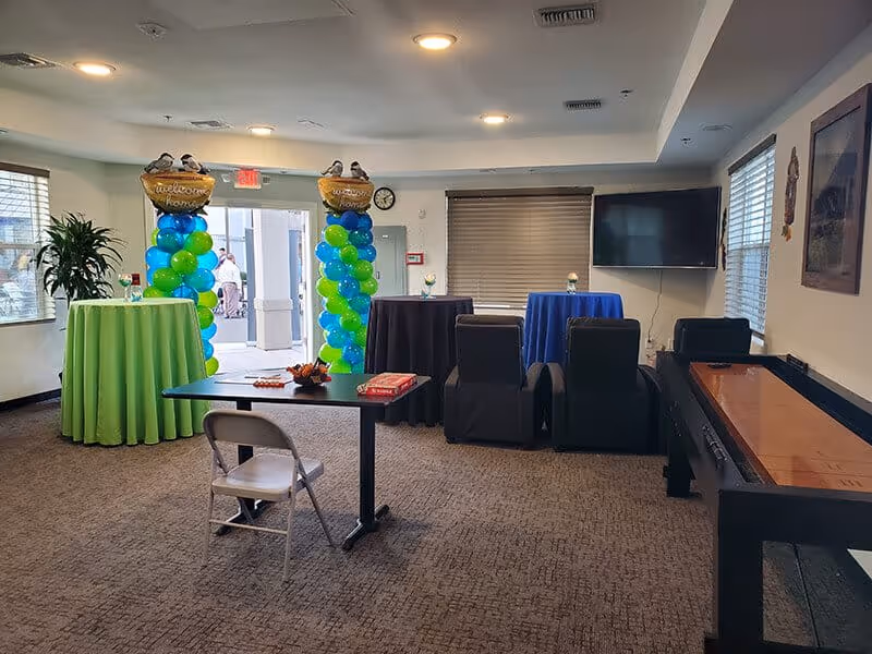 A senior living community activity room with balloon columns, tall covered tables, seating, a TV, and a shuffleboard table.