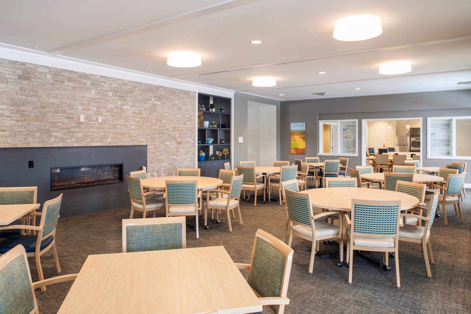 A spacious dining room with multiple round and square wooden tables surrounded by chairs with green upholstery. The room features a modern electric fireplace set into a stone accent wall, built-in shelving with decorative items, and large windows looking into an adjacent room with more tables and chairs. The ceiling has recessed lighting and round light fixtures.
