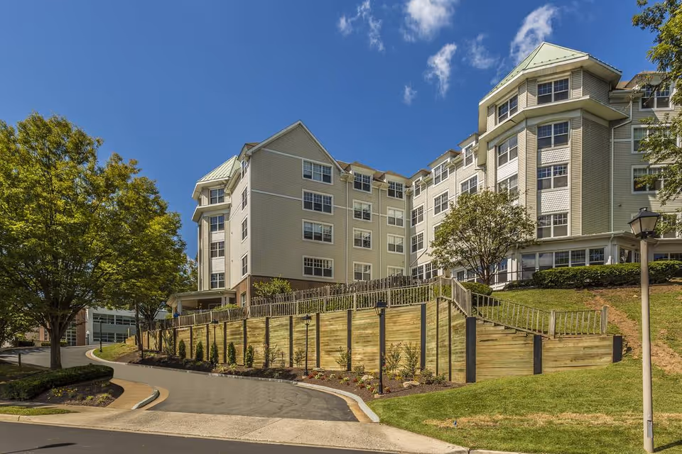 Large multi-story beige senior living building set on a landscaped hillside with a terraced wooden retaining wall and a curved entrance driveway under a blue sky.