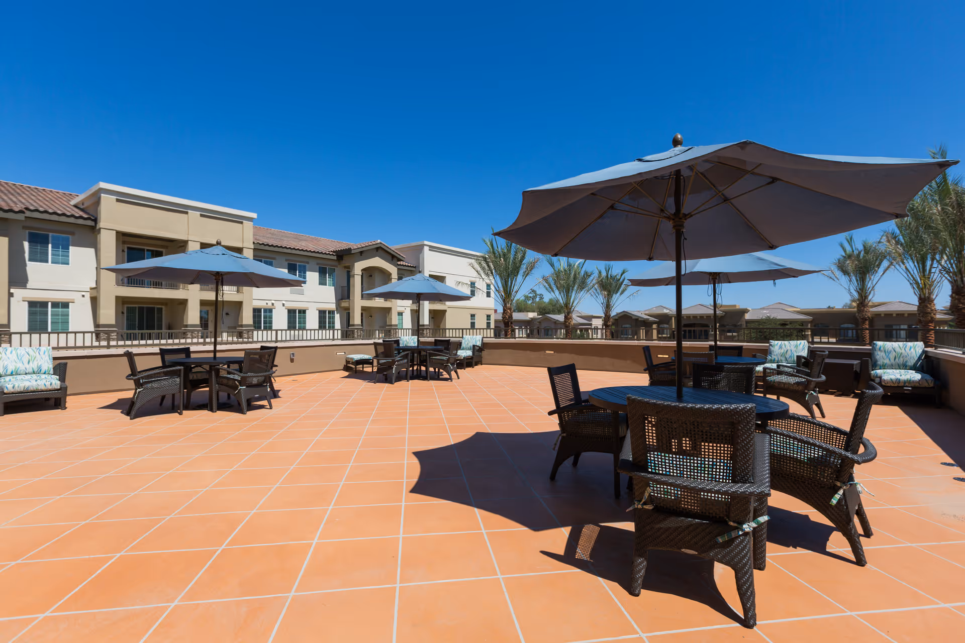 Outdoor patio area with terracotta tiled floor, several black wicker tables and chairs with blue umbrellas providing shade, cushioned lounge chairs along the perimeter, palm trees, and a clear blue sky with a multi-story beige building in the background.