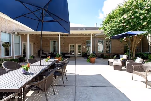Outdoor patio area at Morada Deer Park with multiple seating arrangements including a long table with chairs under a large blue umbrella, and several cushioned wicker chairs and sofas with a smaller blue umbrella. The patio is surrounded by a brick building with windows and doors, and there are potted plants and a tree providing greenery.