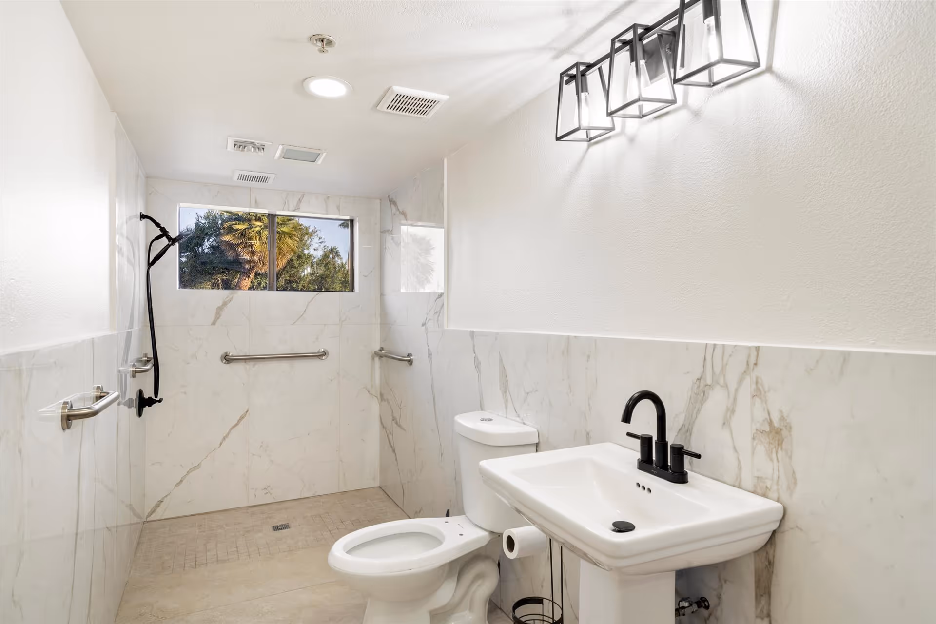 A modern bathroom with white marble walls and beige tiled floor. It features a white toilet, a white pedestal sink with a black faucet, and a walk-in shower area with a black showerhead and grab bars. A window above the shower lets in natural light, and a rectangular light fixture with three bulbs is mounted on the wall above the sink.