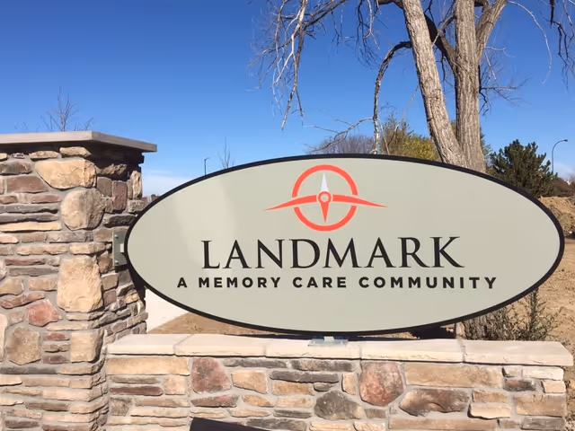 Oval sign reading "Landmark A Memory Care Community" mounted on a stone wall outdoors against a clear blue sky.
