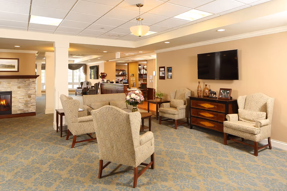 Well-lit common lounge with upholstered armchairs arranged around a coffee table, a wall-mounted TV, and a fireplace in the background.