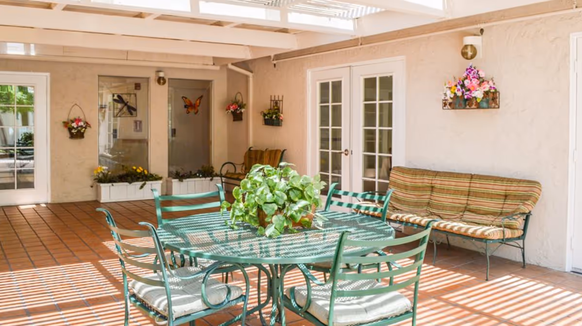 A bright indoor patio area with a green metal table and chairs set, a striped cushioned bench, potted plants on the table and hanging on the walls, and sunlight streaming through a slatted roof creating striped shadows on the terracotta tiled floor.