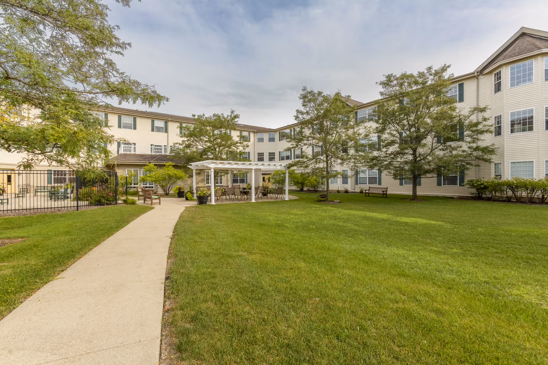 Outdoor courtyard area of a senior living facility with a paved walkway leading to a white pergola with tables and chairs underneath. The courtyard is surrounded by a three-story beige building with many windows. There are several trees, benches, and well-maintained green grass throughout the area.