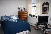 A cozy bedroom with a bed covered in a dark blue comforter and blue patterned pillows. There is a wooden dresser with decorative items on top, a small TV on a stand, and framed pictures on the windowsill and wall. The room has a window with blinds and light-colored walls.