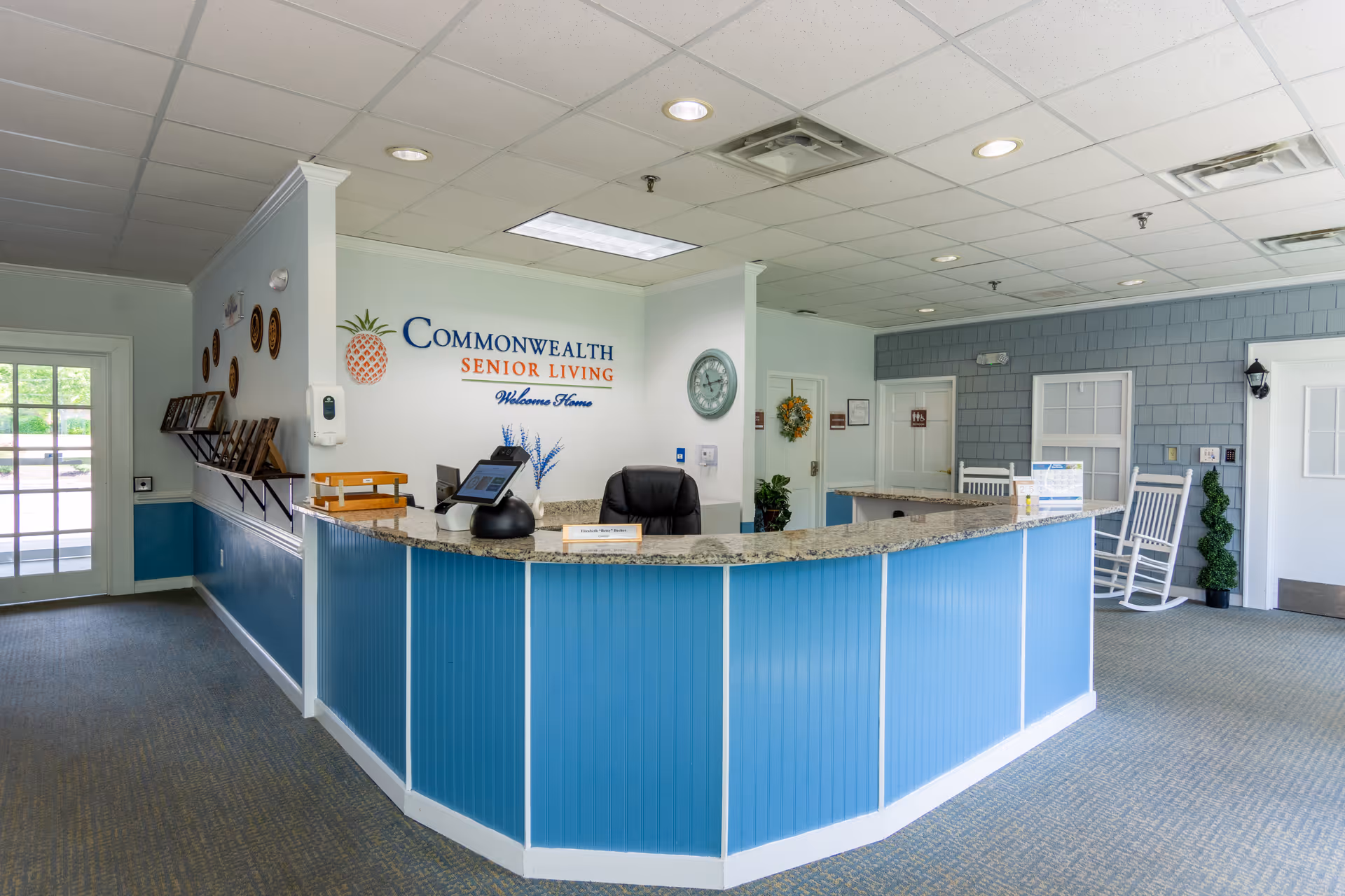 Reception area of a senior living facility featuring a curved blue front desk with a granite countertop, wall sign reading 'Commonwealth Senior Living', and rocking chairs.