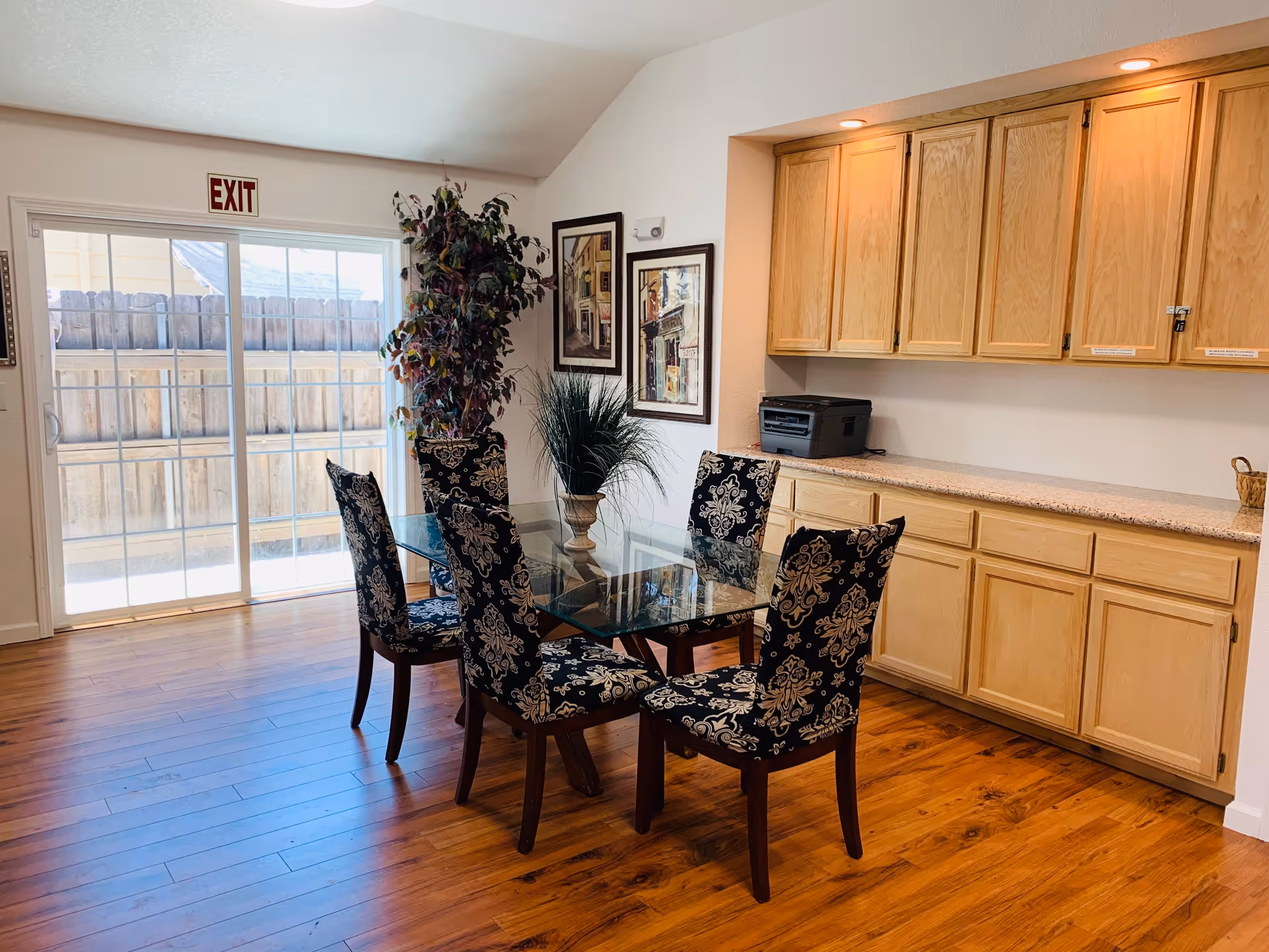 Bright dining area with a glass-top table surrounded by patterned chairs, wooden cabinets and hardwood floors next to a sliding glass door with an EXIT sign.