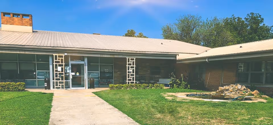 Exterior view of Ruth Wilson Hurley Manor showing a single-story building with a metal roof, large windows, and a central entrance door. There is a concrete walkway leading to the entrance, surrounded by green grass and some landscaping including bushes and a small rock water feature on the right side. Trees are visible in the background under a clear blue sky.