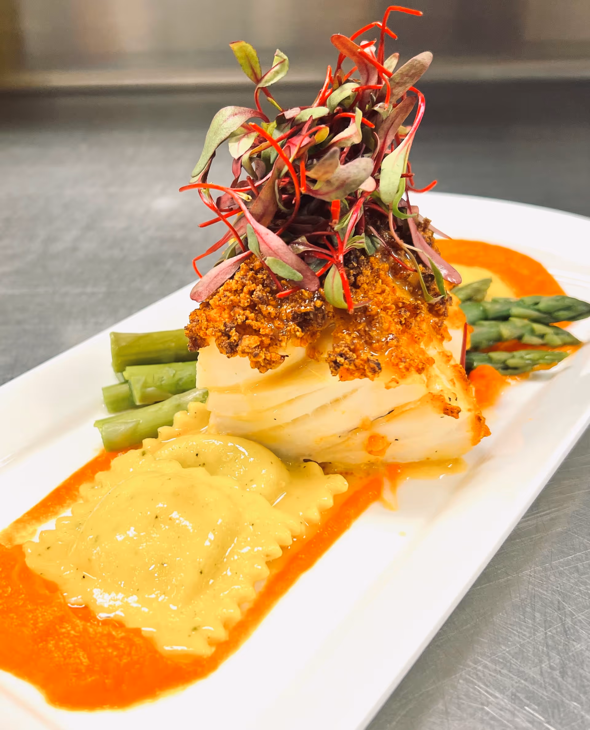 A gourmet plated dish featuring a piece of crusted fish topped with microgreens, served with asparagus spears, ravioli, and a red sauce on a white rectangular plate.