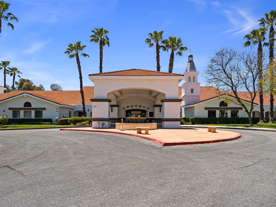 Exterior view of Westmont Village Homes facility featuring a large covered entrance with an archway, surrounded by palm trees and a clear blue sky. The building has a red-tiled roof and white walls with brown trim, and there is a circular driveway in front.