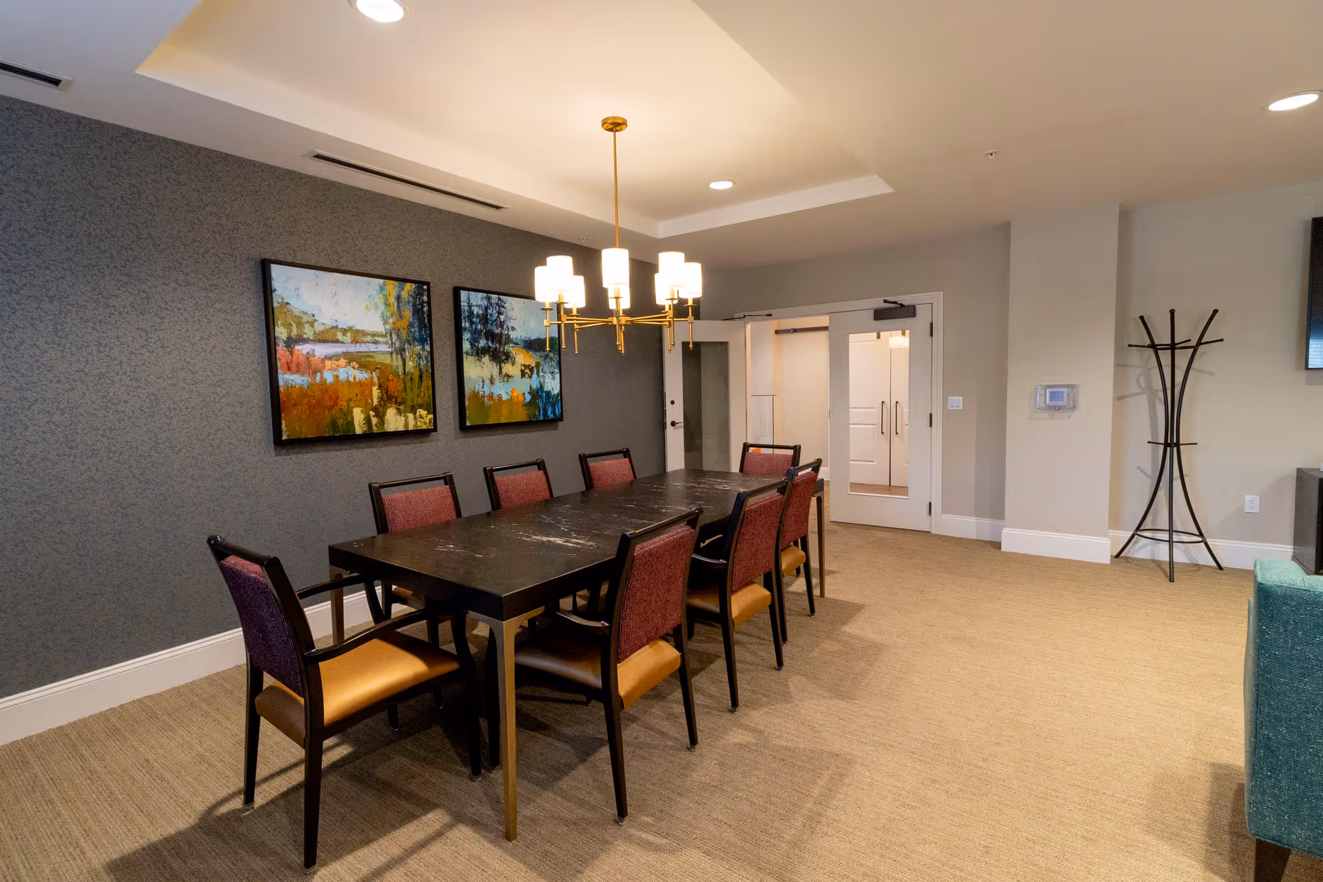 A dining room with a long rectangular black table surrounded by eight chairs with purple backs and tan seats. The room has beige carpet, a gray accent wall with two colorful landscape paintings, a modern chandelier hanging above the table, and a coat rack near a set of white double doors.