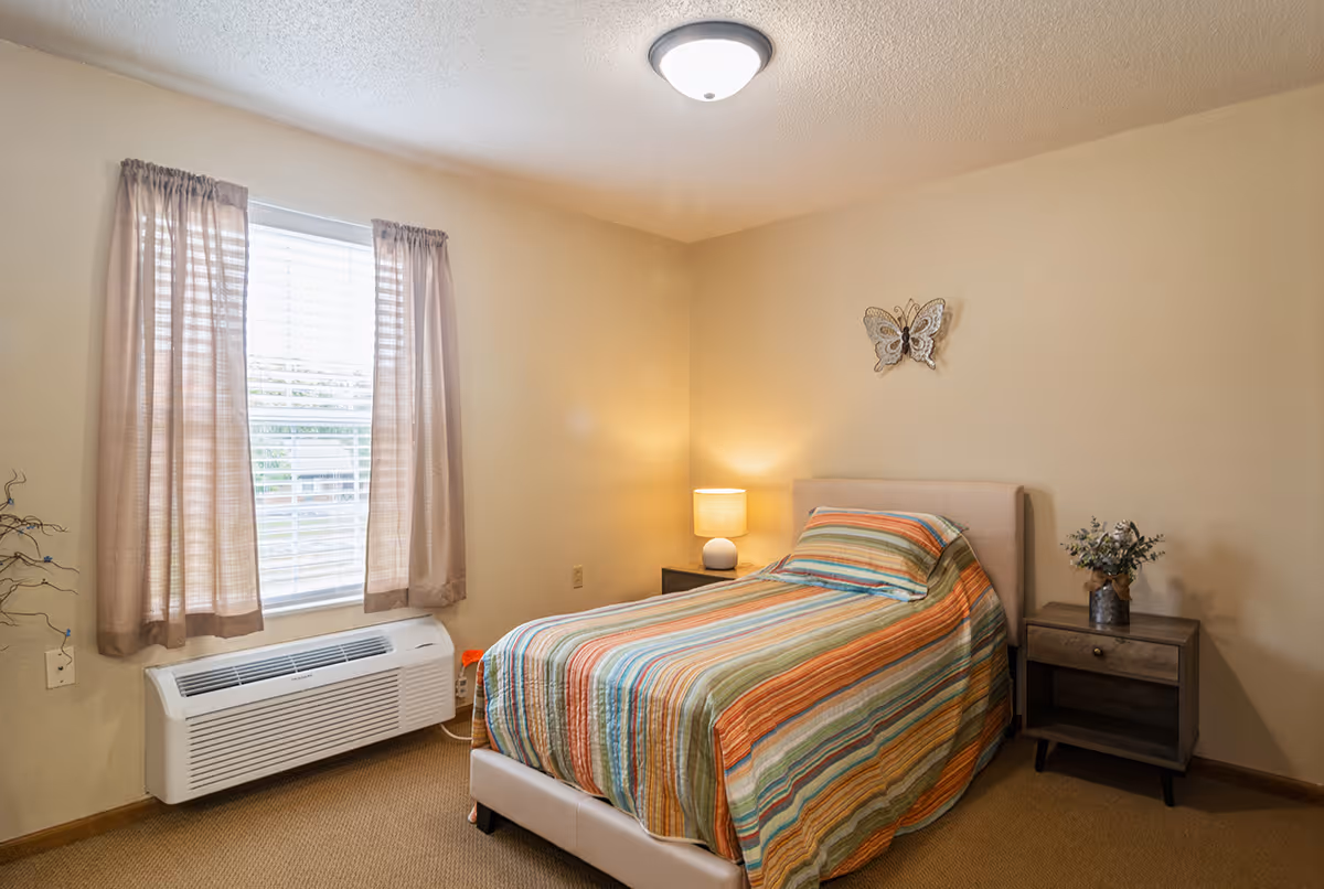 A small bedroom with a single bed covered in a colorful striped bedspread. There is a nightstand with a lamp and a small vase with flowers next to the bed. A butterfly decoration is mounted on the beige wall above the bed. A window with sheer curtains and a white air conditioning unit below it is on the left side of the room.