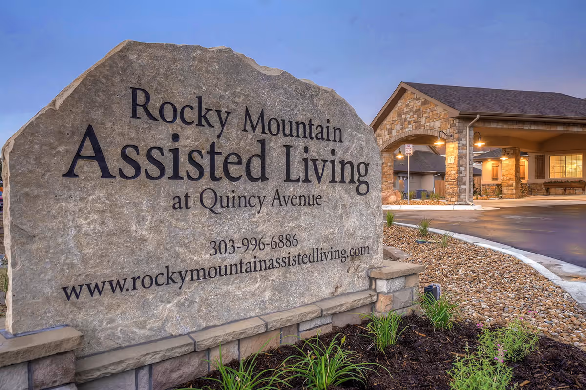 Large stone sign for Rocky Mountain Assisted Living at Quincy Avenue with contact number and website, situated in front of a building entrance with stone pillars and warm lighting during dusk.