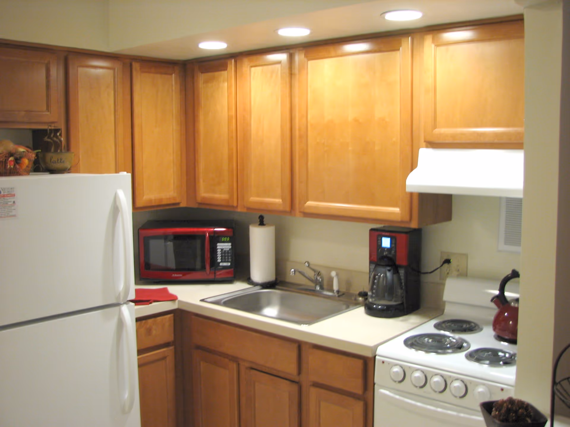 A small kitchen with wooden cabinets, a white refrigerator, a red microwave, a paper towel holder, a coffee maker, a white electric stove with four burners, and a red kettle on the stove. The kitchen has overhead lighting and a stainless steel sink.